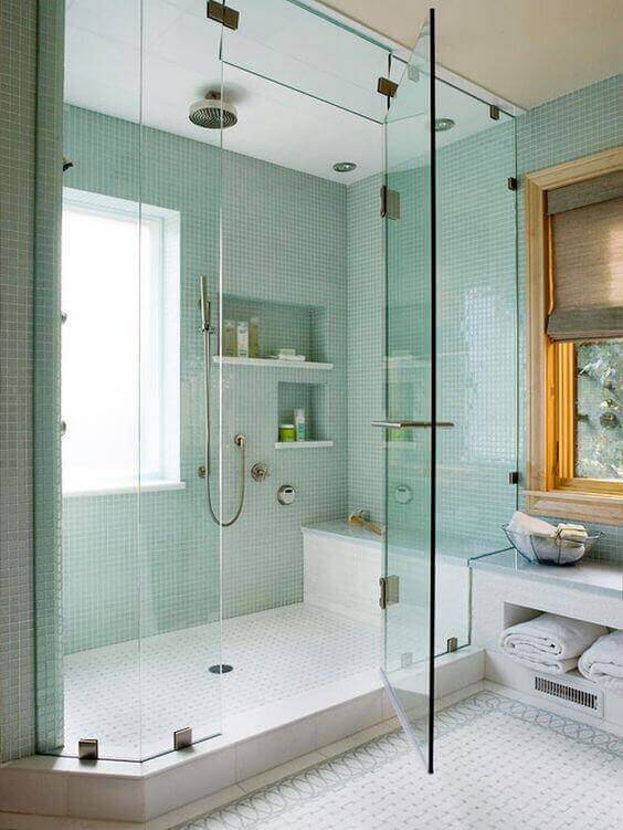 Frameless glass shower enclosure with mint green mosaic tiles, built-in bench, white hexagonal floor tiles, and natural light for a spa-like vibe.