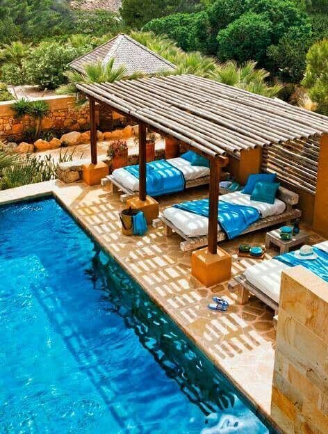 Outdoor lounge area with a wooden pergola over a poolside setting, featuring beige tiles, blue and white lounge chairs, and tropical plants.