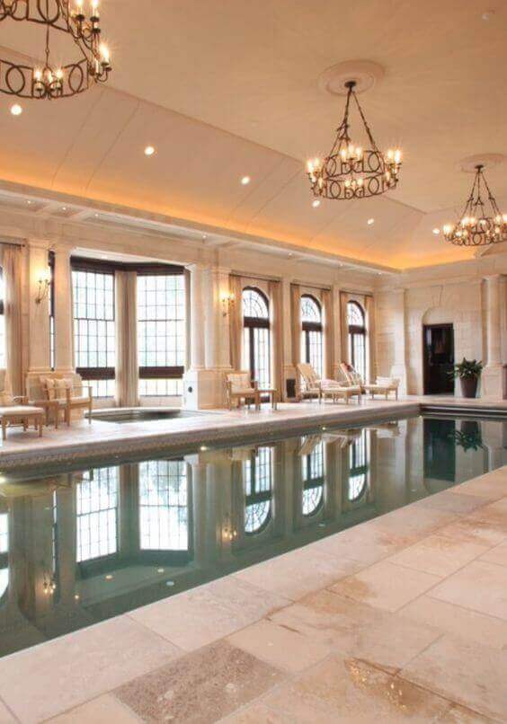 Indoor Swim Spa features a 15x30 ft pool with natural stone tiles, illuminated by chandeliers and large arched windows, creating a tranquil ambiance.