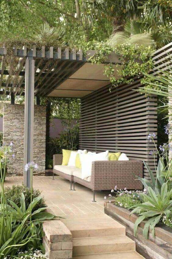 Modern outdoor seating area features a sleek pergola providing partial shade, surrounded by lush greenery and a textured stone accent wall.