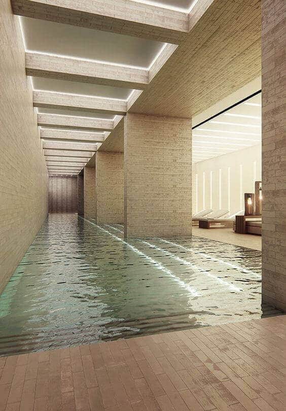 Expansive indoor swim spa with natural light reflection, stone walls, minimalist lighting, and lounge chairs for relaxation.