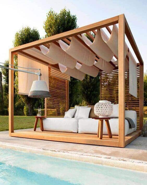 Wooden pergola with plush sectional sofa, light beige drapes, and greenery, creating a cozy outdoor seating area by the pool.