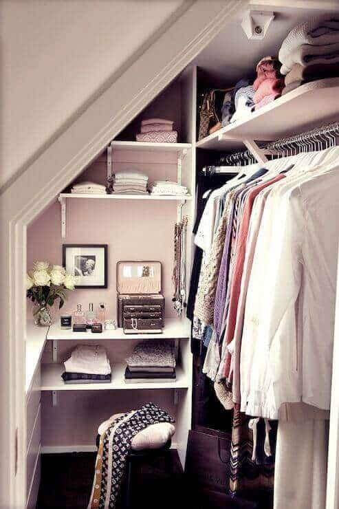Compact attic closet design with white shelving, organized storage for sweaters, and light pink walls, maximizing space elegantly.