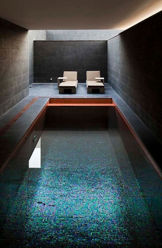Indoor swim spa features dark stone walls and a mosaic tile floor. The 15x6 ft pool is surrounded by modern lounge chairs, creating a spa-like atmosphere.