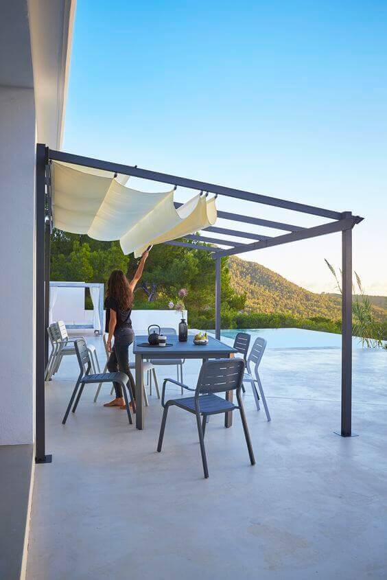 Outdoor patio with a modern pergola featuring a metal frame and retractable canvas cover, surrounded by greenery and mountains.