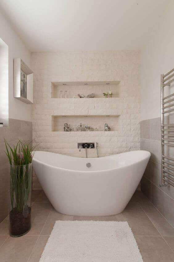 Freestanding white acrylic bathtub in a spacious, minimalist bathroom with light stone walls, natural light, and a tall planter for a spa vibe.