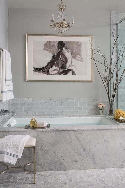 Luxury bathroom with a sleek marble bathtub, glass-enclosed shower, light gray walls, and light blue tile backsplash. Elegant chandelier and cozy towels enhance the spa vibe.