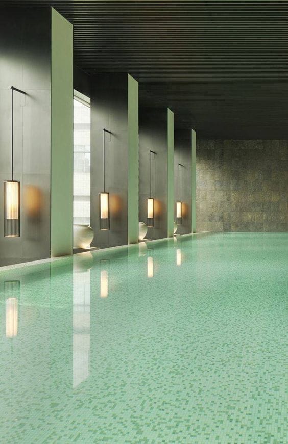 Serene indoor swim spa with an infinity-style layout, glossy surface, muted green tiles, and warm pendant lighting enhancing tranquility.