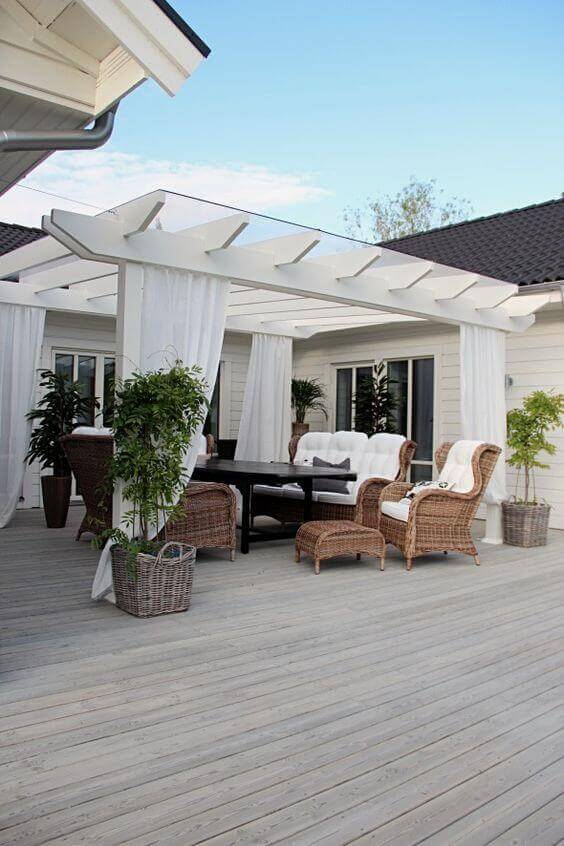 Wooden deck patio features a white pergola, rectangular table with woven chairs, plush seating, and potted plants for greenery.