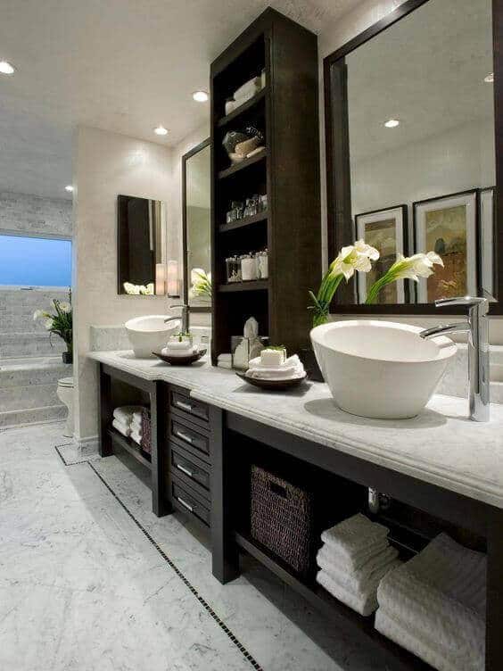 Elegant spa bathroom featuring round vessel sinks on a marble countertop, large mirrors, dark wood storage, and natural light with plant accents.
