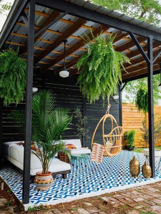 Spacious outdoor patio with patterned blue and white tiles, featuring a wooden roof, black metal beams, and comfortable seating, surrounded by greenery.