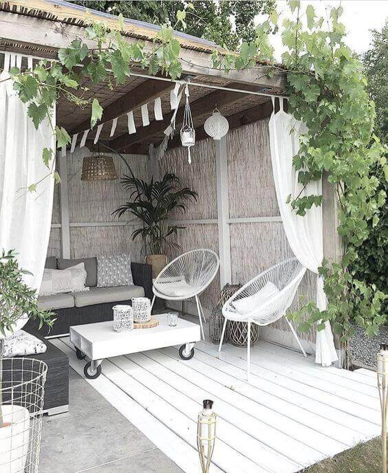 Cozy outdoor lounge under a wooden pergola, featuring a charcoal gray sofa, white Acapulco chairs, and soft ambient lighting.