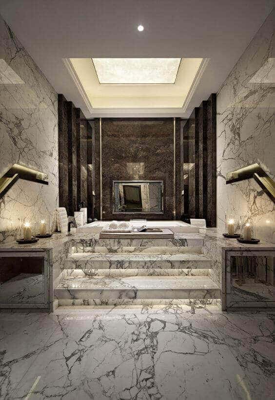 Luxury spa bathroom with large marble tiles, a central bathtub on a stepped platform, ambient candles, dark marble walls, and a skylight.
