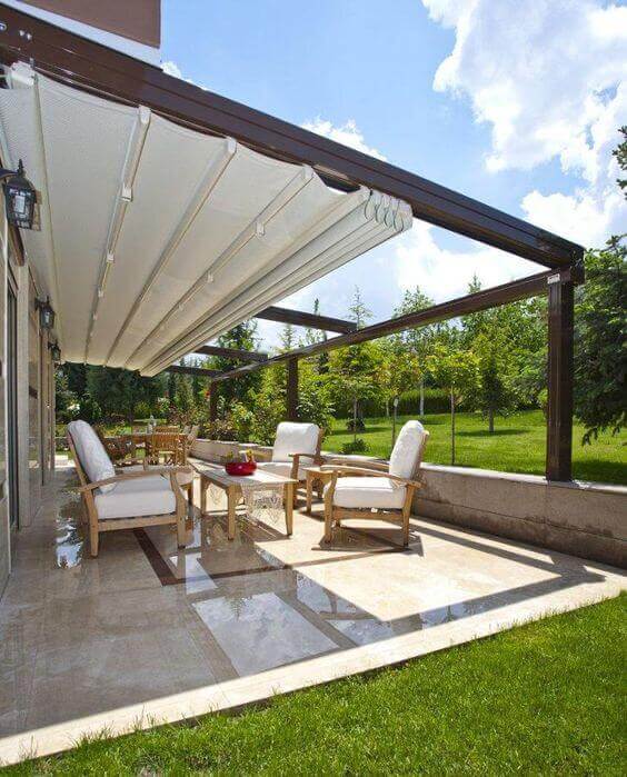 Outdoor living space featuring a large shaded area with a retractable awning, four wooden lounge chairs, and a glass-top table on polished stone tiles.