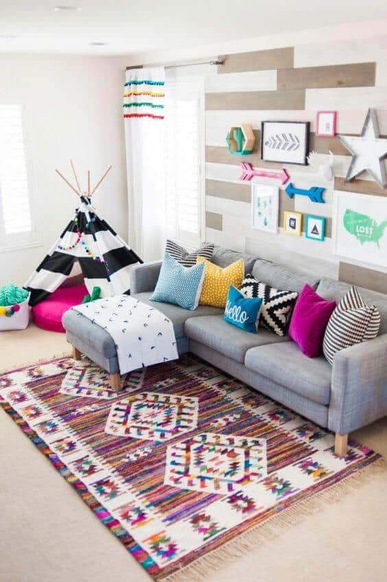 Cozy basement playroom with a gray sofa, colorful pillows, a patterned rug, and a black-and-white striped teepee for kids' creativity.