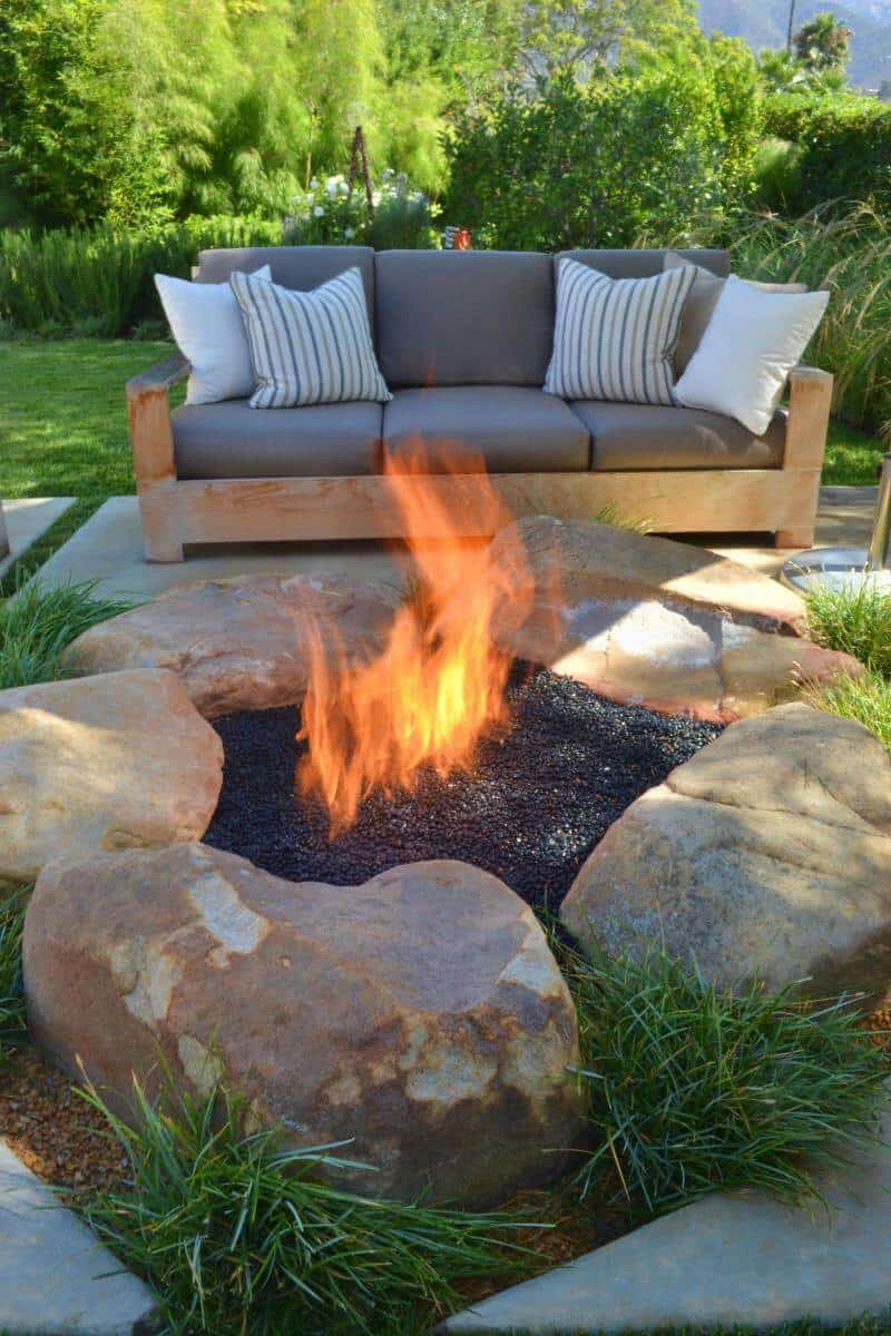 Cozy outdoor living space with a fire pit bordered by smooth stones, black lava rocks, and a modern sectional sofa with gray cushions.