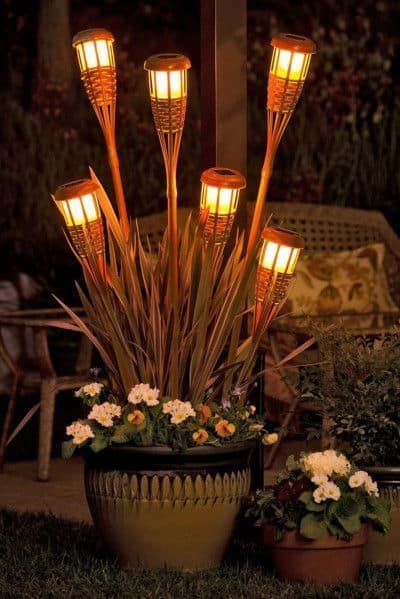 Five decorative lights resembling torches rise from a vibrant 24-inch planter filled with colorful flowers, enhancing backyard ambiance.