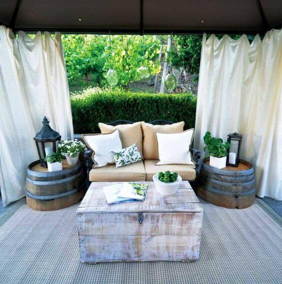 Cozy outdoor seating area with a beige sofa, lush greenery, and upcycled wooden barrels as side tables, perfect for budget-friendly gatherings.