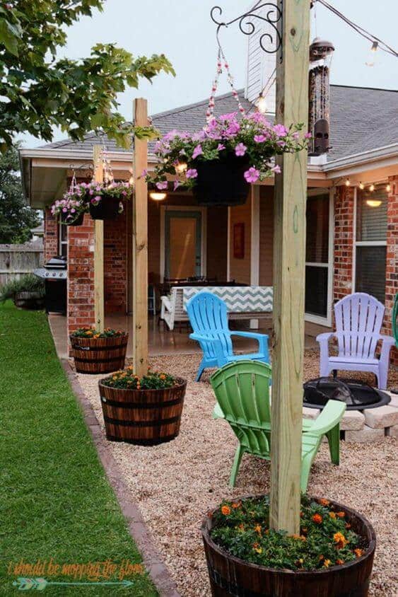 Cozy backyard with Adirondack chairs around a fire pit, wooden posts with pink flower planters, and gravel ground bordered by lush grass.