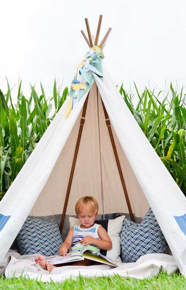 Cozy kids play tent made of soft white fabric and plush cushions, set in a grassy area surrounded by tall corn plants.