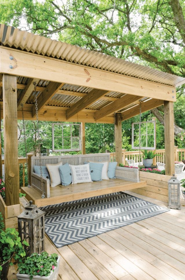 Cozy small backyard deck featuring a wooden platform, pergola with metal roofing, swing seat, chevron rug, and lush greenery for a relaxing atmosphere.