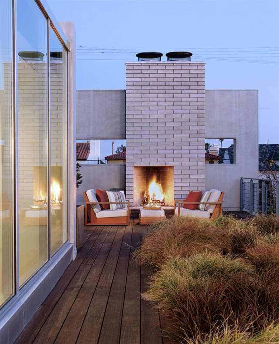 Outdoor patio with a central gray brick fireplace, modern lounge chairs, dark wooden planks, and ornamental grasses, perfect for relaxation.
