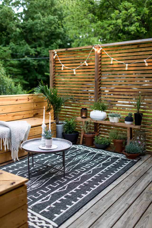 Cozy small backyard deck featuring wooden benches, a round metal coffee table, and potted plants, illuminated by string lights.