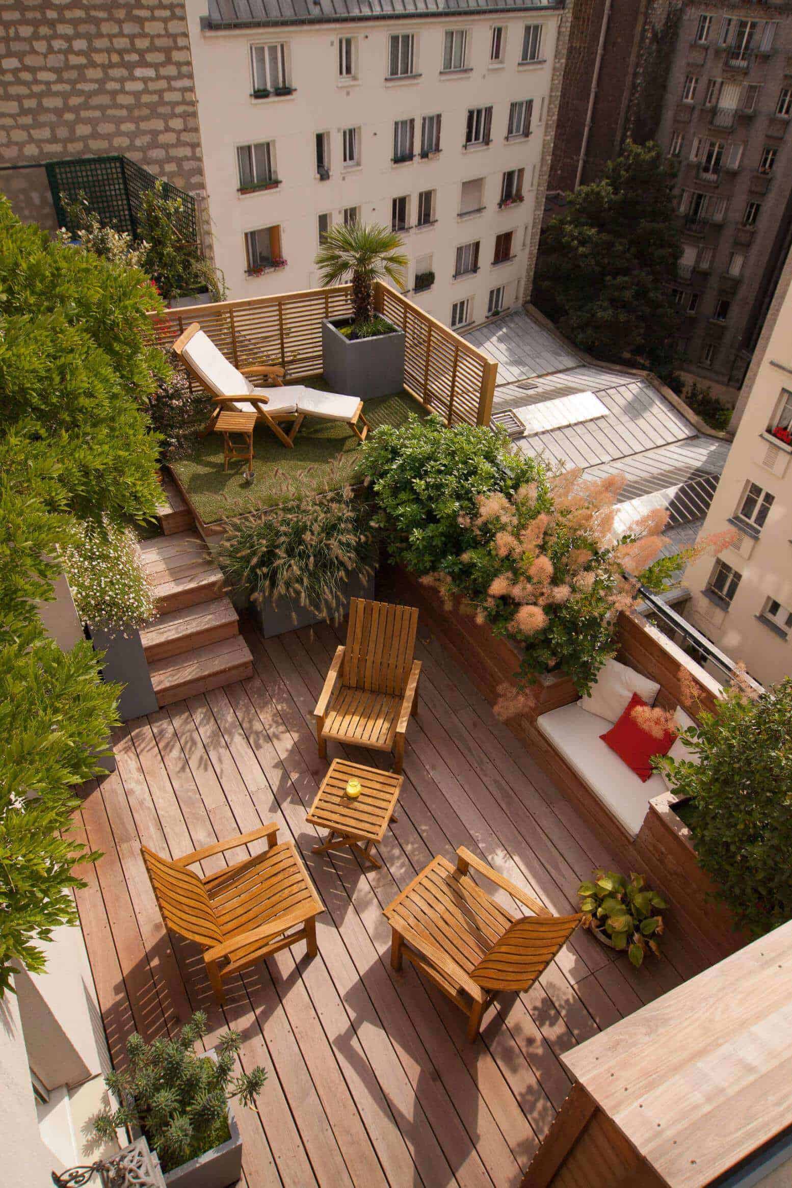 Outdoor terrace with a wooden deck, four slatted chairs around a coffee table, a lounge chair, and potted shrubs for an inviting atmosphere.
