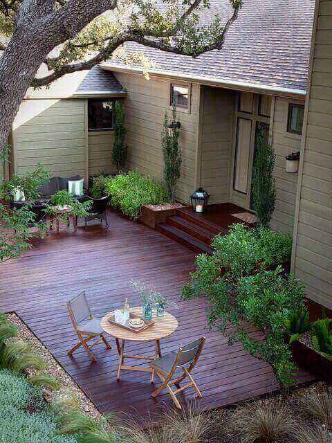 Cozy small backyard deck with a stained wooden surface, circular table, three folding chairs, and surrounded by lush green plants.