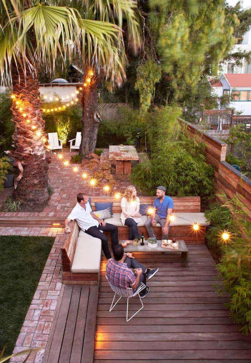 Cozy outdoor living space features a wooden deck with brick pathways, sectional seating for five, and string lights, ideal for gatherings.