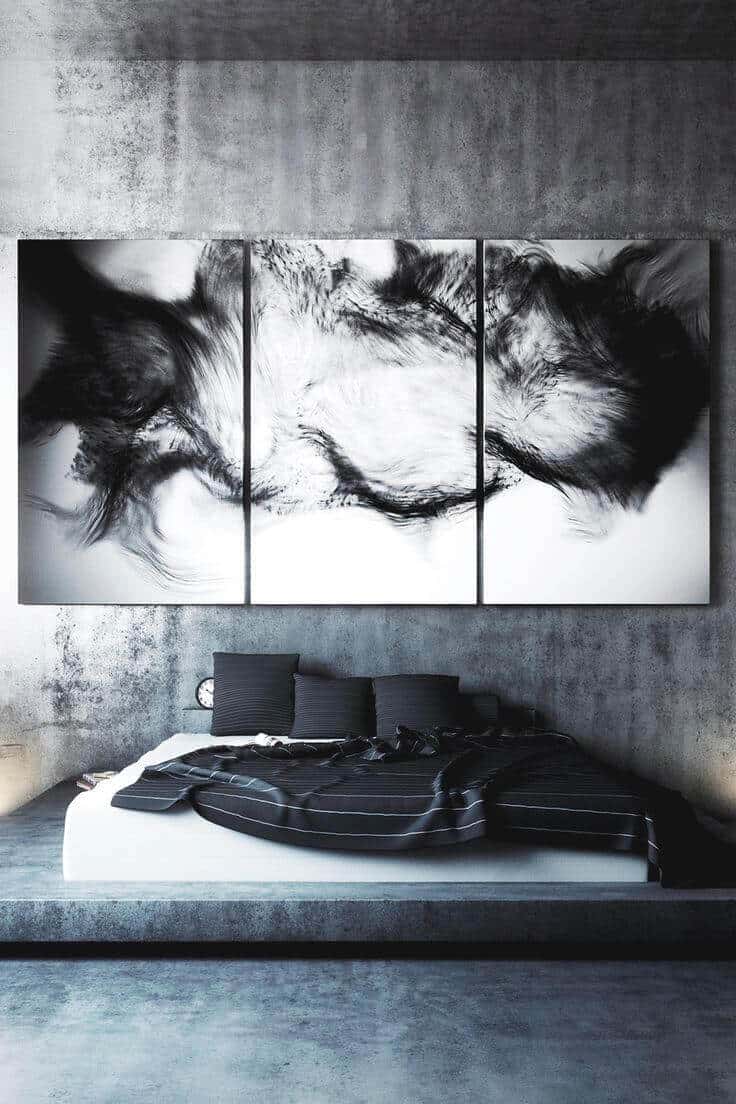 Minimalist grey bedroom featuring a low-profile platform bed with dark bedding. A striking black-and-white triptych decorates the concrete wall, enhancing the serene atmosphere.