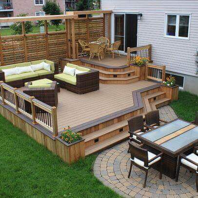 Multi-level outdoor deck with composite wooden planks, featuring wicker sofas, a circular dining table, and integrated planters.
