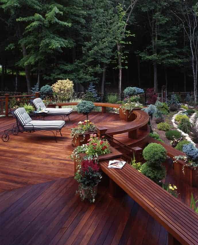 Curved seating areas with cushioned lounge chairs and vibrant planters on a dark-stained wood deck, surrounded by lush trees for privacy.