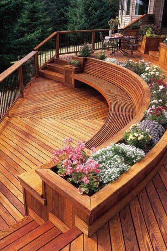Curved wooden deck made from treated hardwood with integrated seating, surrounded by colorful flower beds and lush trees for privacy.
