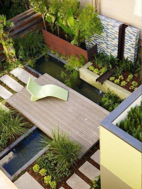 Wooden deck in a modern garden design, 10x8 feet, surrounded by water features and greenery. Includes a sleek lounge chair and stepping stones.