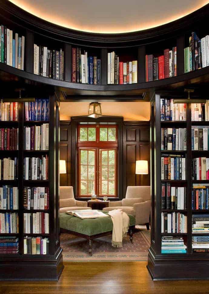 Cozy reading nook with dark wood bookshelves, two neutral armchairs, a green ottoman, and warm lighting, perfect for a serene reading experience.