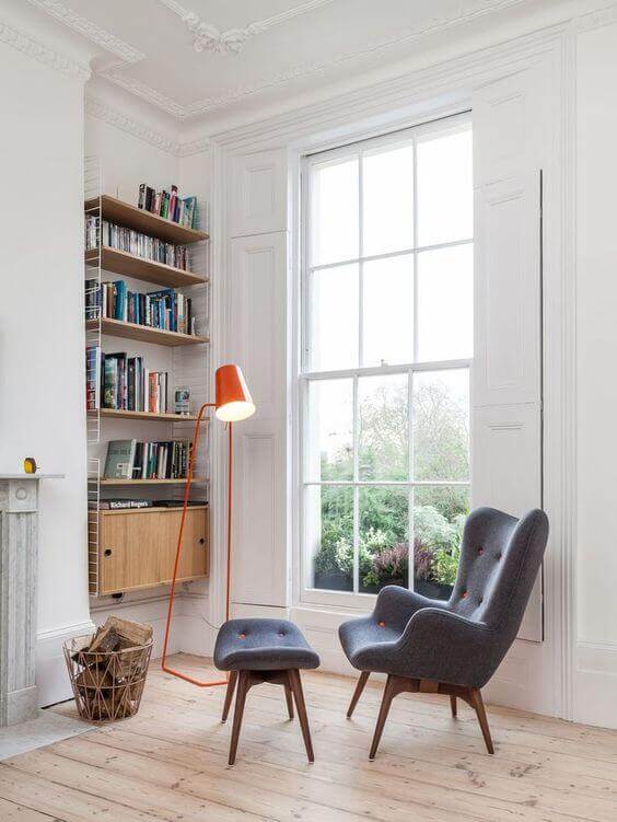 Cozy reading nook with a gray upholstered armchair and footstool, wooden bookshelf, and stylish floor lamp, bathed in natural light.