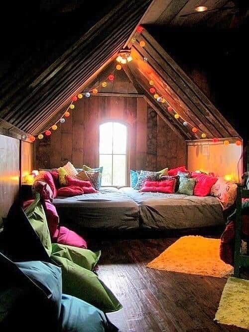 Attic bedroom with a sloped wooden ceiling, colorful cushions, a large plush bed, and ambient lighting, creating a cozy reading nook atmosphere.