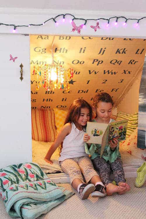 Cozy kids reading nook with alphabet wallpaper, oversized cushion, colorful pillows, and a whimsical chandelier, creating a warm retreat.