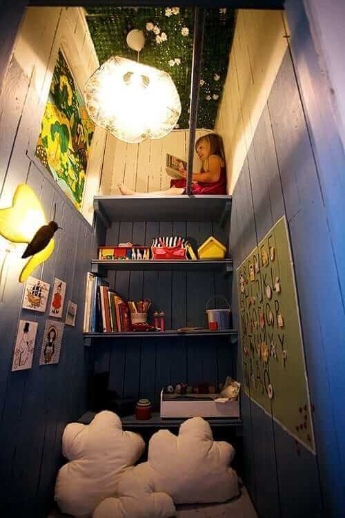 Cozy kids reading nook with blue wooden walls, a shelf for books and art supplies, plush cloud cushions, and whimsical ceiling light.