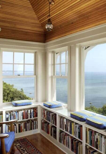 Cozy reading nook with built-in bookshelves, large windows for ocean views, natural wood ceiling, and inviting blue and yellow cushions.