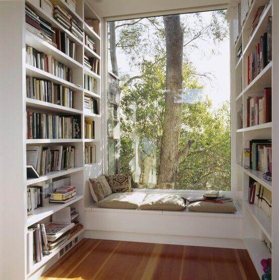 Cozy reading nook featuring built-in white shelves filled with books, a cushioned bench with pillows, and a large window overlooking lush trees.