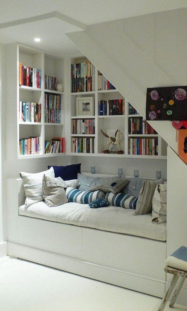 Cozy reading nook under a staircase, featuring a built-in bench with cushions, custom white shelving filled with books, and warm LED lighting.