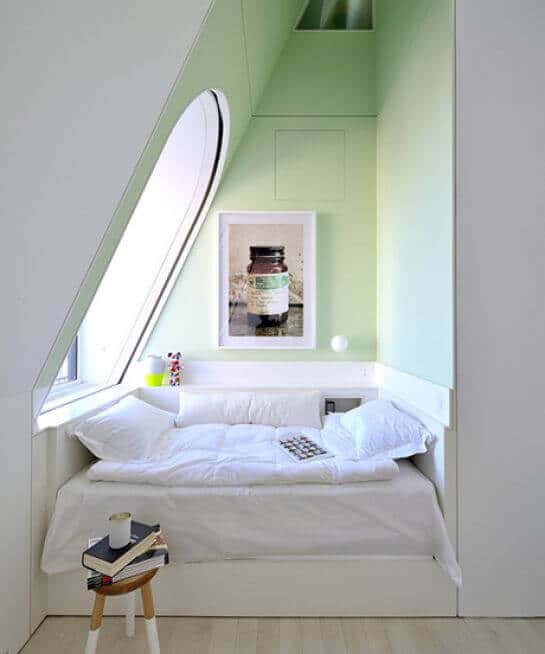 Cozy reading nook under a sloped ceiling, featuring a built-in bed with white bedding, light green walls, and a large window for natural light.