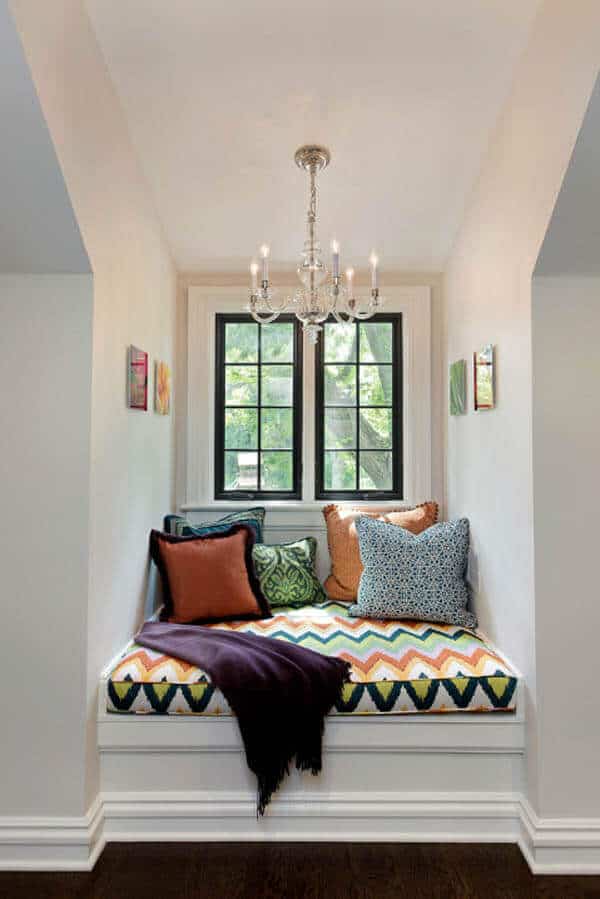 Cozy reading nook with a window seat, vibrant zigzag cushion, decorative pillows, and natural light from black-framed windows.
