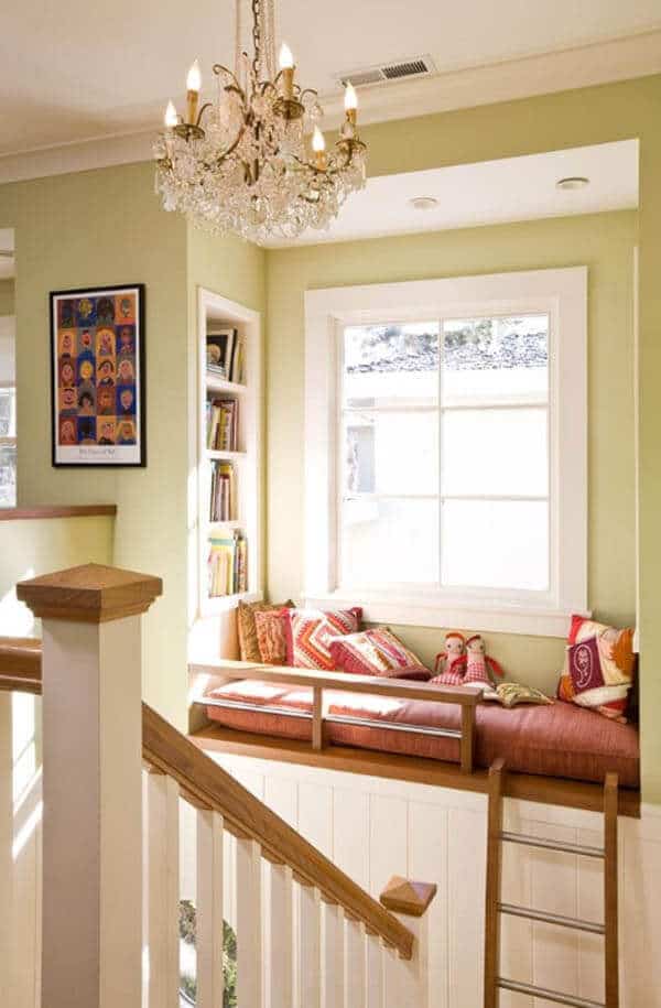 Cozy reading nook with a built-in window seat, soft green walls, vibrant cushions, and a crystal chandelier, surrounded by bookshelves.