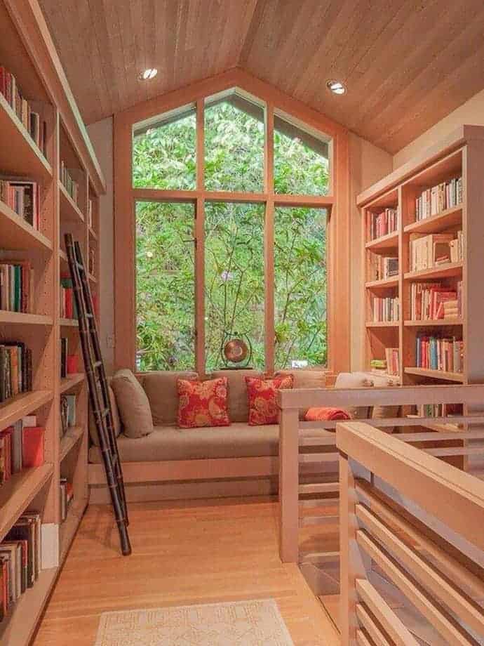 Cozy reading nook with light hardwood bookshelves, a neutral sofa with red pillows, and a desk by large windows, blending style and comfort.
