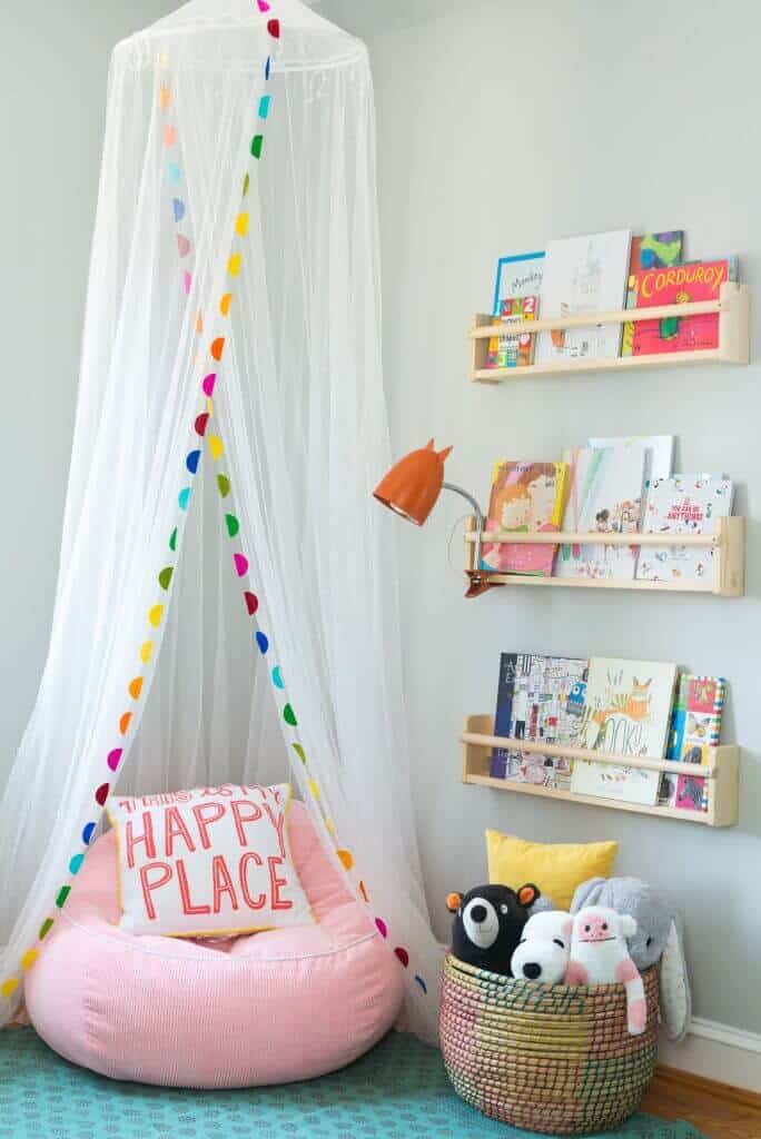 Cozy kids reading nook with a pink bean bag chair, 'HAPPY PLACE' pillow, white canopy, colorful pom-poms, and wooden bookshelves.