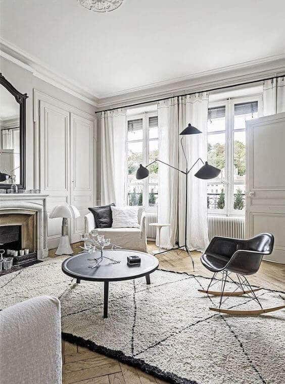 27 Contemporary Living Room Ideas for a Modern Haven Space 23 Minimalist contemporary living room with a neutral palette, featuring a round black coffee table, modern rocking chair, and elegant armchair. Large windows enhance the airy feel, complemented by subtle molding and a vintage fireplace.