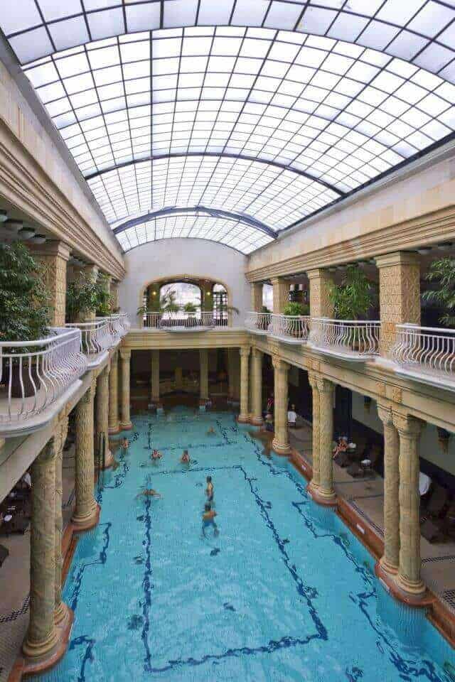 Indoor pool with a large rectangular area, surrounded by ornate stone columns and a skylight ceiling, featuring lush greenery and elegant tiles.